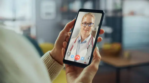 Photo of Close Up of a Female Chatting in a Video Call with Her Family Doctor on Smartphone from Living Room. Ill-Feeling Woman Making a Call from Home with Physician Over the Internet.