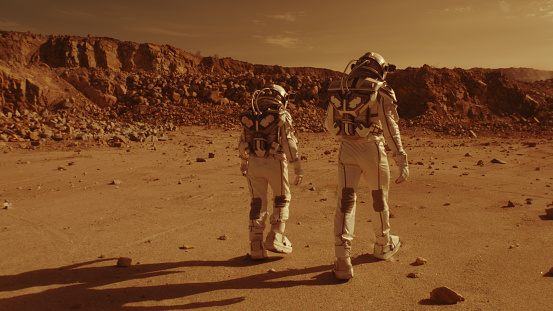 Tracking shot of anonymous people in spacesuits walking on arid ground together during colonization of Mars