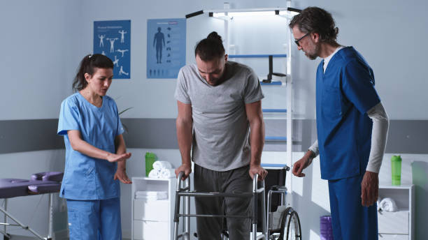 Handicapped man trying to stroll with walking frame near doctors Female and male therapist helping adult patient on wheelchair to stroll with walking frame during rehabilitation session in clinic walking aide stock pictures, royalty-free photos & images