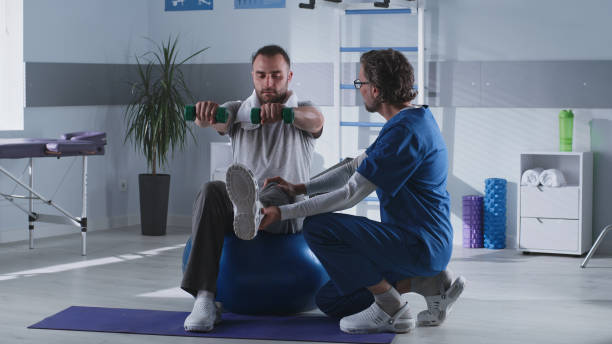 Mature therapist supporting male patient on fit ball Middle aged man in medical uniform supporting adult male patient with dumbbells sitting on fit ball and exercising in rehabilitation clinic physical therapy stock pictures, royalty-free photos & images