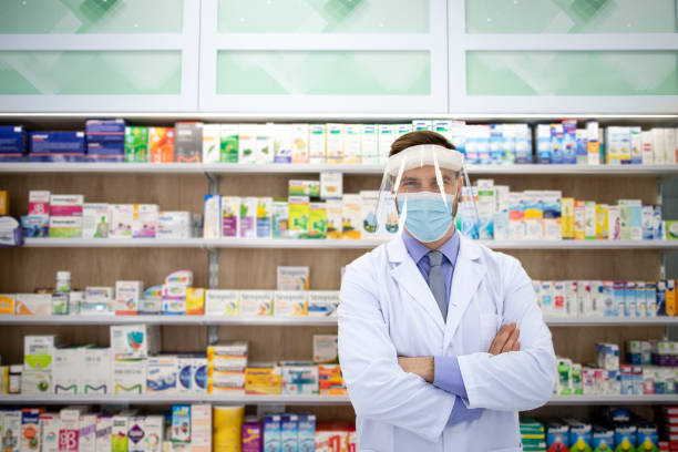 retrato de farmacéutico con escudo facial y bata blanca de pie en la farmacia durante la pandemia de coronavirus. - pill bottle pharmacy medicine shelf fotografías e imágenes de stock