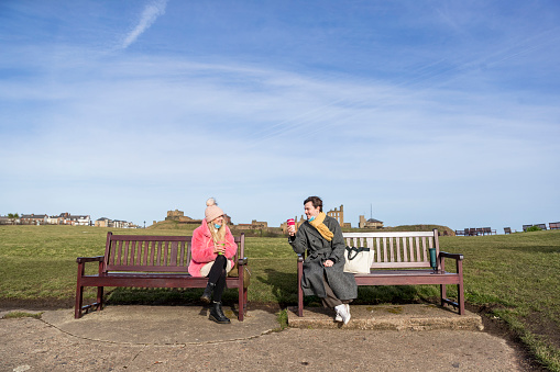 Couple sitting on benches social distancing drinking hot drinks in the North East of England during the Covid pandemic. They are dressed warmly and are wearing protective face masks.
