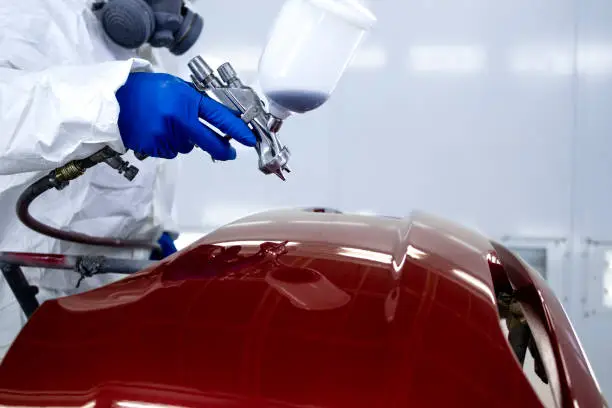 Photo of Car painter in protective clothes and mask painting automobile bumper with metallic paint and varnish in chamber workshop.