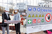 A warning board attached to the site fence at a construction site. In the background, an architect or civil engineer is talking to a woman