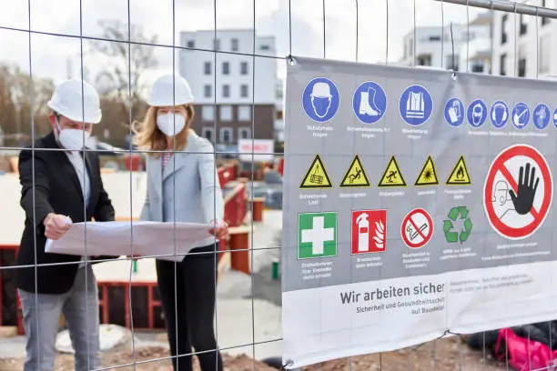 Photo of A warning board attached to the site fence at a construction site. In the background, an architect or civil engineer is talking to a woman