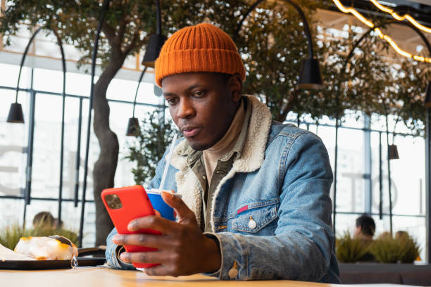 istock Black man in knitted hat and jacket uses smartphone in cafe 1324688067