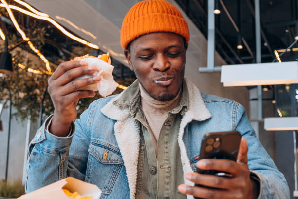 African-American guy eats tasty hamburger looking into phone Positive African-American guy in orange hat eats tasty hamburger looking into mobile phone at table in decorated cafe closeup fast food restaurant stock pictures, royalty-free photos & images
