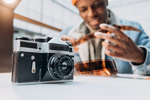 Positive African-American guy photographer in denim jacket checks vintage film sitting at table focus on old camera closeup