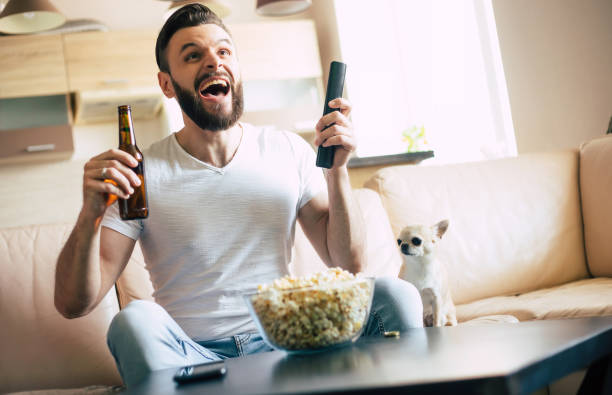 istock Handsome young excited modern bearded man is relaxing on the couch while watching football match on TV 1324688338