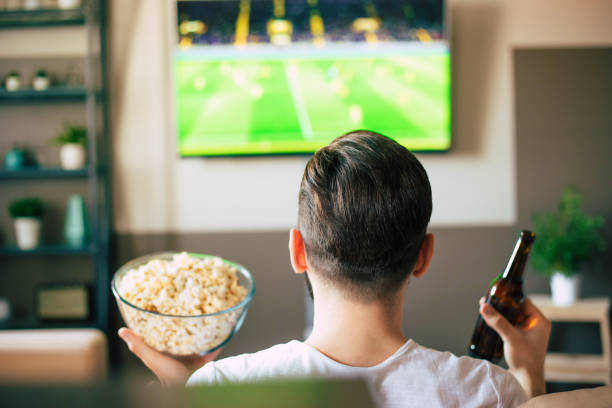 istock Handsome young excited modern bearded man is relaxing on the couch while watching football match on TV 1324689129