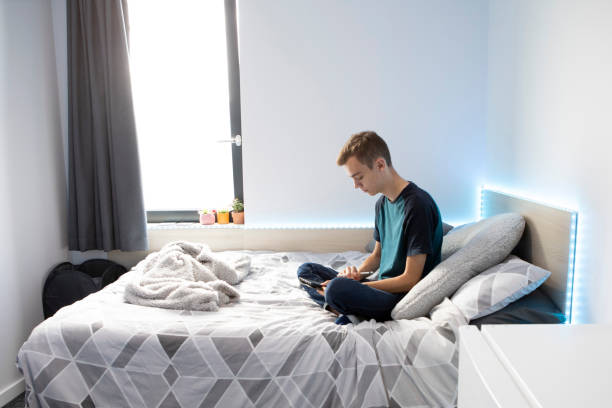 istock Young male student sitting on a bed using a stylus pen and digital tablet. He is wearing a turquoise t-shirt and denim jeans and there are lights around the bed. 1324690234