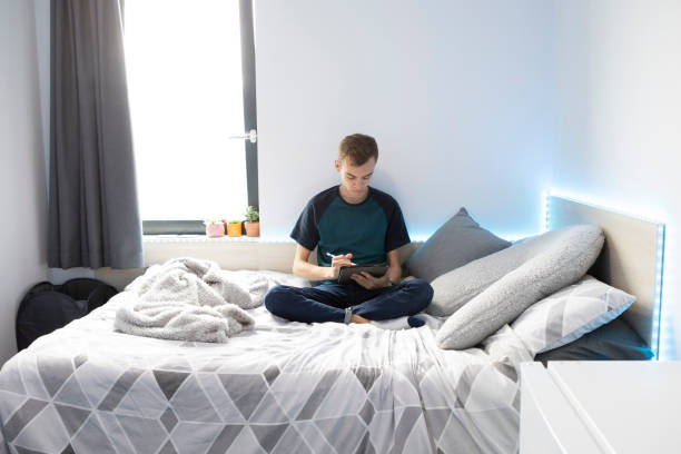 istock Young male student sitting on a bed using a stylus pen and digital tablet. He is wearing a turquoise t-shirt and denim jeans and there are lights around the bed. 1324690261