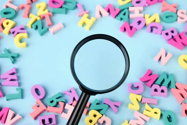 Photo of Lots of multicolored wooden letters scattered on a blue background. Magnifying glass in center.