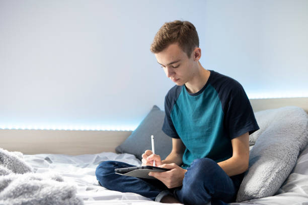 istock Young male student sitting on a bed using a stylus pen and digital tablet. He is wearing a turquoise t-shirt and denim jeans and there are lights around the bed. 1324690318