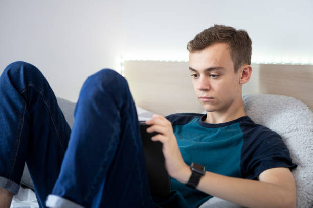 istock Young male student sitting on a bed using a digital tablet. He is wearing a turquoise t-shirt and denim jeans and there are lights around the bed. 1324690320