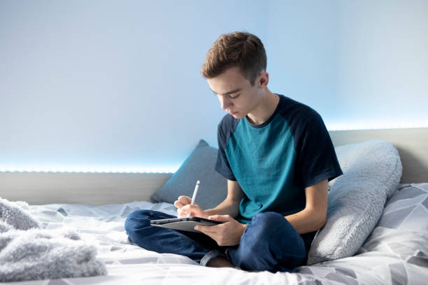istock Young male student sitting on a bed using a stylus pen and digital tablet. He is wearing a turquoise t-shirt and denim jeans and there are lights around the bed. 1324690848