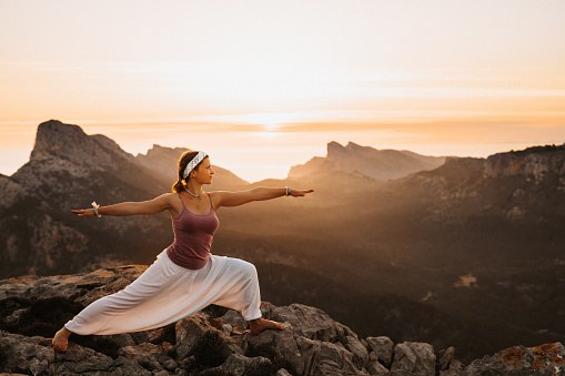 Beautiful girl doing the yoga warrior position during the sunrise in a mountainous landscape in Majorca. Color editing with added grain. Part of a series.