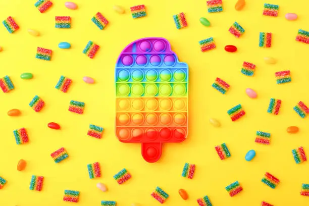 Photo of Rainbow Pop it fidget toy next to the colorful marmalade candies on yellow background.