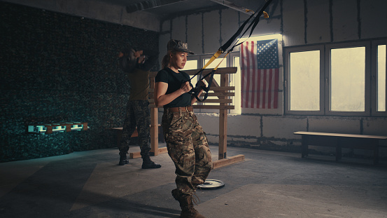 View of tired woman in camouflage pants doing lunges near squadmate during training in dark gym of USA military base