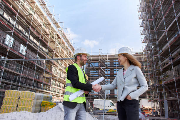 un arquitecto o ingeniero civil con documentos de planificación se reúne con un cliente o colega en el gran sitio de construcción y ambos usan cascos protectores blancos. - hardhat construction men handshake fotografías e imágenes de stock