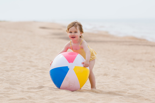 Happy little girl playing with colorful big ball on sand at sea beach in warm sunny summer day. Looking at camera. Front view.
