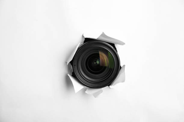 istock Hidden camera lens through torn hole in white paper 1324700196