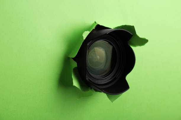 istock Hidden camera lens through torn hole in green paper 1324700531