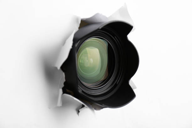 istock Hidden camera lens through torn hole in white paper 1324700538