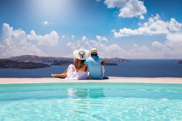 una elegante pareja con ropa de verano se sienta junto a la piscina y disfruta de la vista al mar mediterráneo - honeymoon fotografías e imágenes de stock