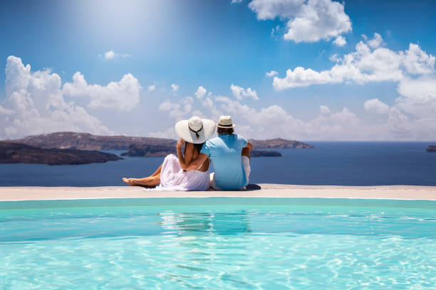 A elegant couple in summer clothes sits by the pool and enjoys the view to the mediterranean sea A elegant couple in summer clothes sits by the pool and enjoys the view to the mediterranean sea in Greece during their summer holidays santorin stock pictures, royalty-free photos & images