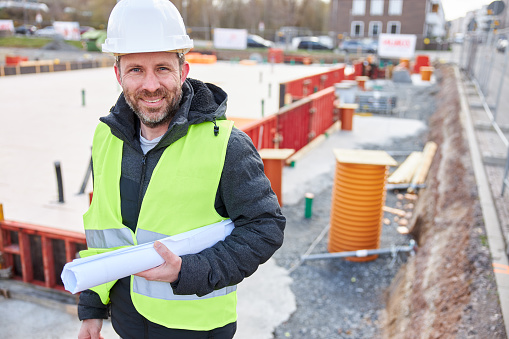 An architect or civil engineer stands with rolled up planning documents and a safety vest in front of a foundation under construction on the construction site and wears a white hard hat. Photographed in high resolution with copy space