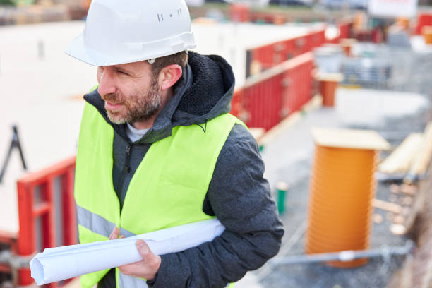istock An architect or civil engineer stands with rolled up planning documents and a safety vest in front of a foundation under construction on the construction site and wears a white hard hat. 1324704098