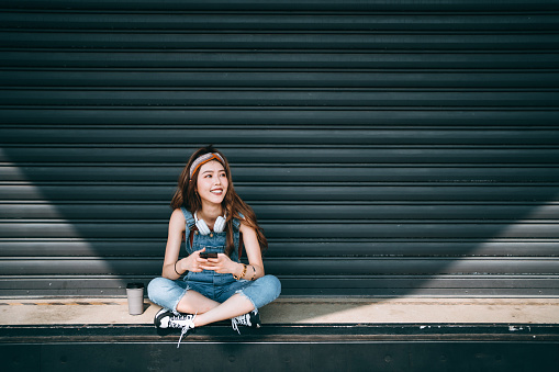 Smiling Asian female university student wearing headphones around her neck, looking up while text messaging on smartphone, sitting in front of roller shutter in school campus against sunlight. Teenage lifestyle and technology