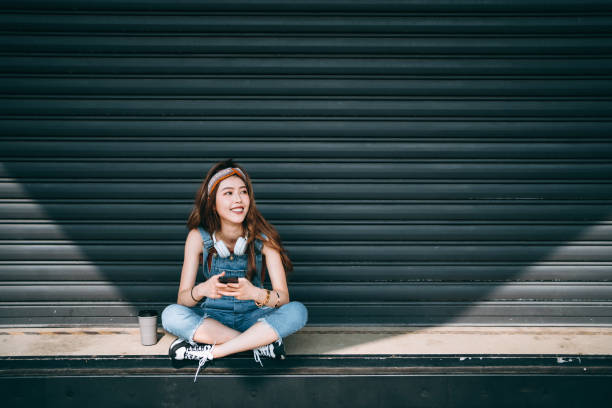 smiling asian female university student wearing headphones around her neck, looking up while text messaging on smartphone, sitting in front of roller shutter in school campus against sunlight. teenage lifestyle and technology - chinese ethnicity audio imagens e fotografias de stock