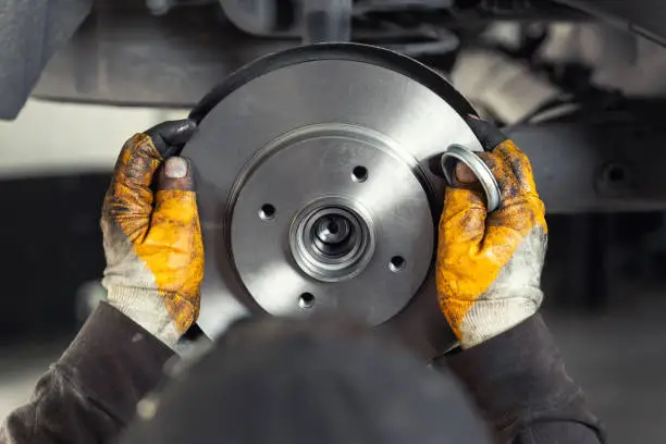 Closeup male tehnician mechanic greasy hands in gloves install new car oem brake steel rotor disk during service at automotive workshop auto center. Vehicle safety checkup and maintenance concpet.