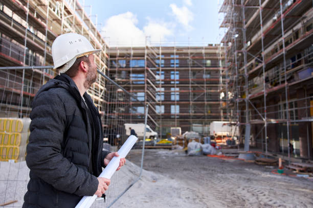 istock An architect or civil engineer stands with rolled up planning documents in front of a scaffolded apartment building and wears a white hard hat. 1324705171