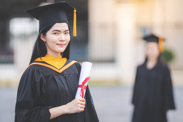 A young Asian woman university graduate in graduation gown and mortarboard holds a degree certificate stands in front of the university building after participating in college commencement stock photo