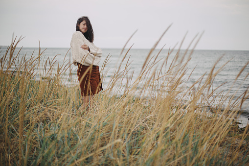 Beautiful stylish woman with windy hair in knitted sweater posing among wild grass. Carefree moment. Fashionable young female standing on windy coast. Authentic and Tranquility. Copy space