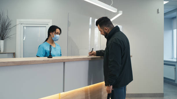 Male patient signing contract on clinic reception Bearded man speaking with Asian female receptionist in mask and signing contract the walking away while visiting modern medical center real time stock pictures, royalty-free photos & images