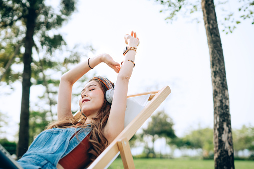 Beautiful, carefree young Asian woman with her eyes closed relaxing on deck chair in park, listening to music with headphones and hands raised. Enjoying Summer days outdoors. Music, teenage lifestyle and technology