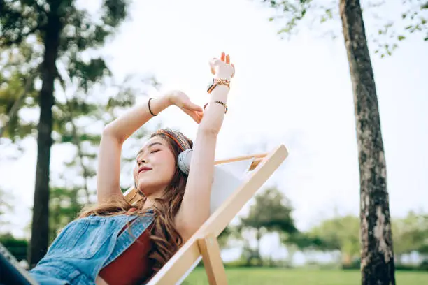 Photo of Beautiful, carefree young Asian woman with her eyes closed relaxing on deck chair in park, listening to music with headphones and hands raised. Enjoying Summer days outdoors. Music, teenage lifestyle and technology