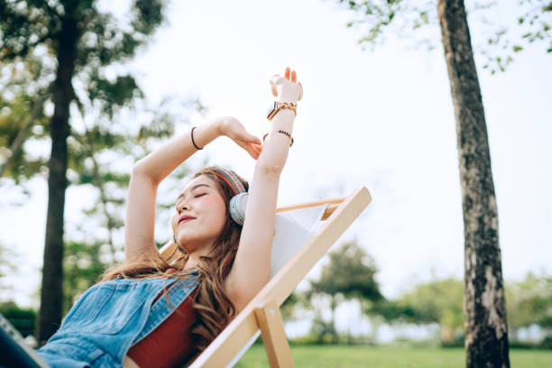 beautiful, carefree young asian woman with her eyes closed relaxing on deck chair in park, listening to music with headphones and hands raised. enjoying summer days outdoors. music, teenage lifestyle and technology - music listening women relaxation imagens e fotografias de stock