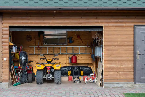 Photo of Facade front view open door ATV quad bike motorcycle parking messy garage building with wooden siding at home driveway backyard and lawn path. House warehouse for tools and equipment . Garage sale