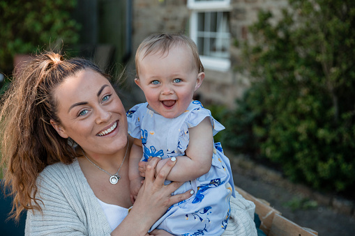 A front-view shot of a young caucasian mother holding her young baby girl, they are smiling and looking cheeky at the camera, they both have beautiful blue eyes.