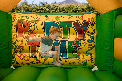 A shot of a young caucasian boy having fun bouncing on a bouncy castle at a birthday party in a backyard.