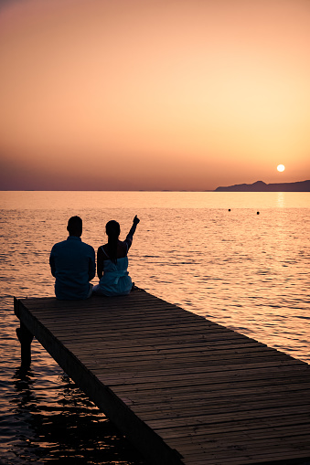 a couple seated on a wooden jetty, looking at colorful sunset on the sea, men, and women watching a sunset in Crete Greece Europe