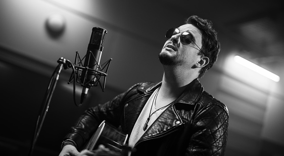 Adult male rocker playing acoustic guitar and singing on microphone in recording studio. Music, arts and entertainment concept.