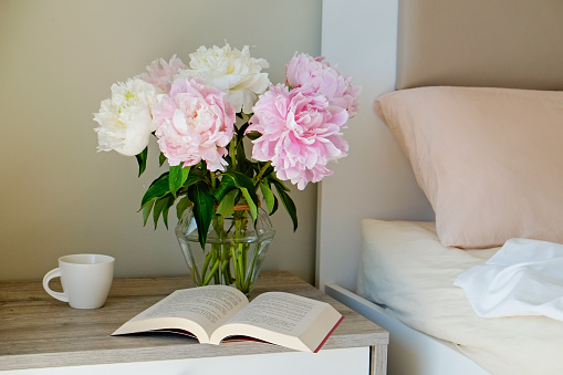 Close up shot of the nightstand with a book and bouquet of peonies in a glass vase near the unmade bed. Good morning concept. Bedroom full of natural light. Copy space for text, background, top view.