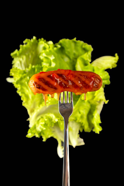 Grilled sausage on a fork, lettuce leaves in the background. Isolated on a black background. Cholesterol, junk food. Selective focus.