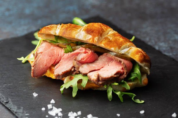 Fresh baked croissant sandwich with arugula and roast beef on a black slate board stock photo
